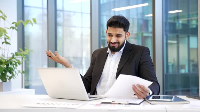 Confused frustrated businessman having difficulty with paperwork sitting at workplace in business office. Puzzled male worker in suit looks at documents and laptop screen and can't understand problem.