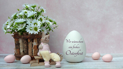 Grußkarte Frohe Ostern:  Osterhase mit rosa Ostereiern vor einem Blumenstrauß sitzend. Auf einem Osterei steht der Text wir wünschen ein frohes Osterfest. © Racamani