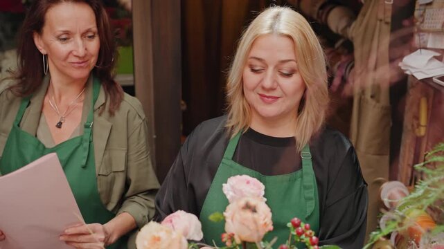Caucasian florist arranging peach rose bouquet on counter. Cozy boutique scene with mentor guiding apprentice, green apron, pastel peonies and hypericum berries, hands trimming stems, scissors