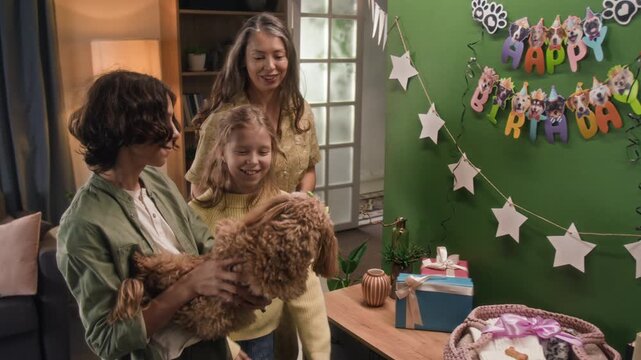 Positive family of two teenagers and mother pointing at funny garland on green wall showing it to brown-coloured moodle dog celebrating birthday