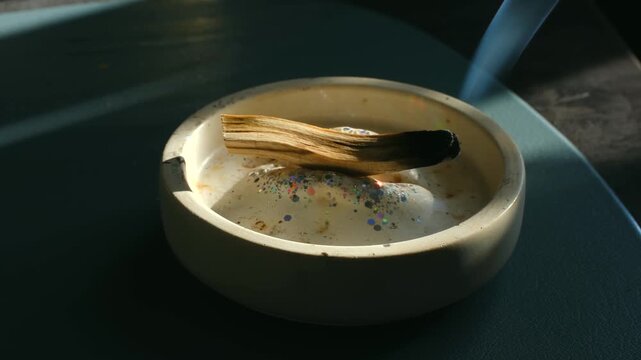 Burning palo santo sacred wood stick smoldering in handmade ceramic bowl with glitter. The incense is used in spiritual rituals for cleansing, purification, and creating a positive atmosphere