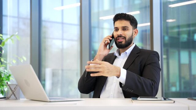 Businessman talking on mobile phone sitting at work desk at workplace in office. Confident manager in formal suit communicates with client or has conversation with business partner using smartphone.