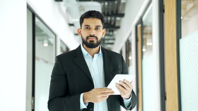 Portrait of serious businessman in formal suit using a digital tablet and looking at camera in modern business center hallway. Male employee reading message, browsing, chatting online with a client. 