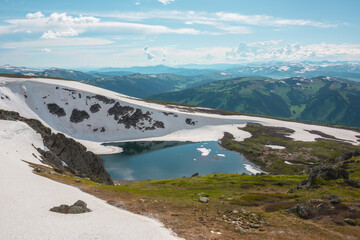 Scenic sunlit landscape with alpine lake in rocky snowy cirque near stone hill top during thaw. Ice floats in mountain lake among rocks and snows with view to forest mountain range under cloudy sky. © Daniil