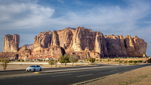 Sandstone rock formation in Al Ula, with steep vertical cliffs and rugged textures, desert terrain with sparse shrubs and paved road, modern car passing under partly cloudy sky. Aula Saudi Arabia