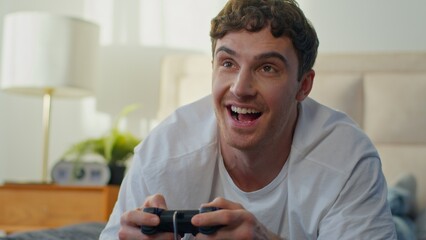 Happy Caucasian young man lying bed at home playing video game joystick smiling guy enjoying console gaming fun hobby male hands holding controller technology virtual cyberspace entertainment bedroom © Yuliia