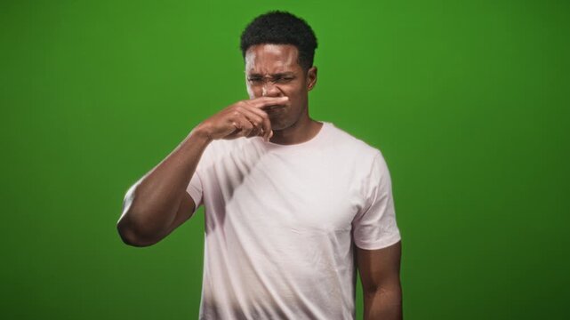 Man pinches nose in studio with green backdrop and pink tshirt, grimacing at a foul smell; disgust discomfort.