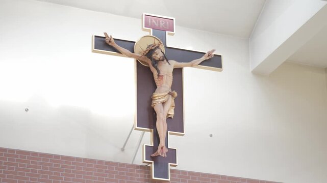 A wooden crucifix of Jesus Christ hanging on a white church wall above a brick pattern. Low angle static shot
