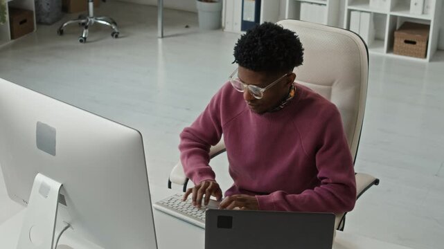 High angle medium shot of young adult African American programmer typing on desktop computer at workstation with laptop while fixing errors in code at modern office