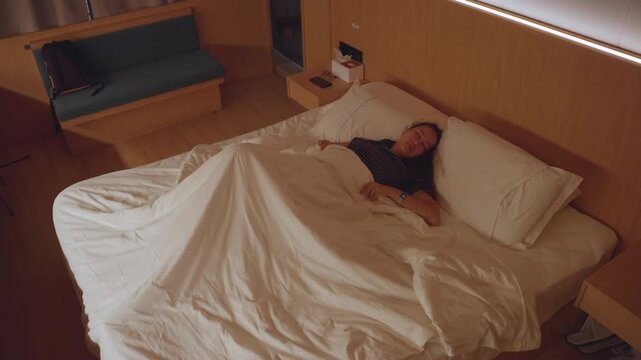 Hotel bed restless female guest tossing, white duvet in disarray, dim bedside lamp glow, turning under covers, insomnia and anxiety, overhead perspective, boutique hotel room, nighttime ambiance, solo