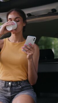 Happy African American woman drinking coffee in car trunk using mobile phone smiling female girl relaxing smartphone browsing social media gadget automobile vehicle transport businesswoman outdoors