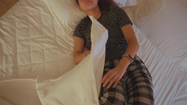 Hotel bed black woman lying under sheets, overhead view shows curly hair on pillow, bedside table with phone and lamp, warm ambient lighting, pensive mood, solo traveler resting after long journey