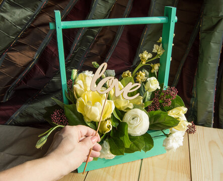 Women's hand holding wooden topper with word Love, box with spring flowers