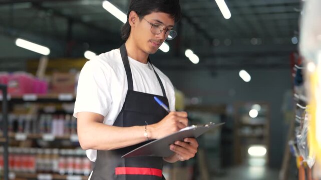 Young male employee checking stock and taking notes on a clipboard in a hardware store aisle