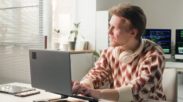 Medium shot of young adult male software developer wearing headphones over neck typing on laptop at working desk then squinting eyes while looking at sunlight during workday at office