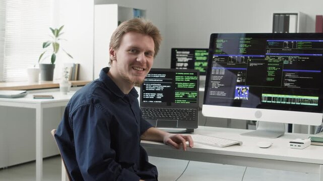 Portrait of young adult male IT specialist typing on computer keyboard editing code then turning to camera with smile sitting at desk in modern office