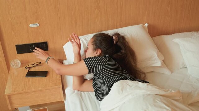 Hotel bed night restless female guest reaching for phone then hugging pillow, tossing and turning across sheets, lamp glow shifting to darkness as scene moves from wakeful reach to drifting sleep,