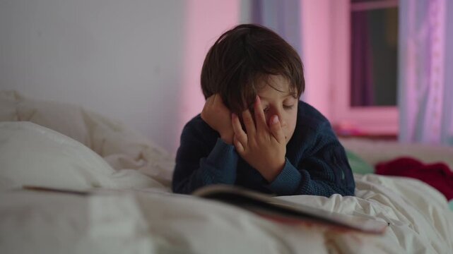 Young boy lying on bed reading book with focused expression during quiet evening at home showing curiosity and imagination in cozy bedroom
