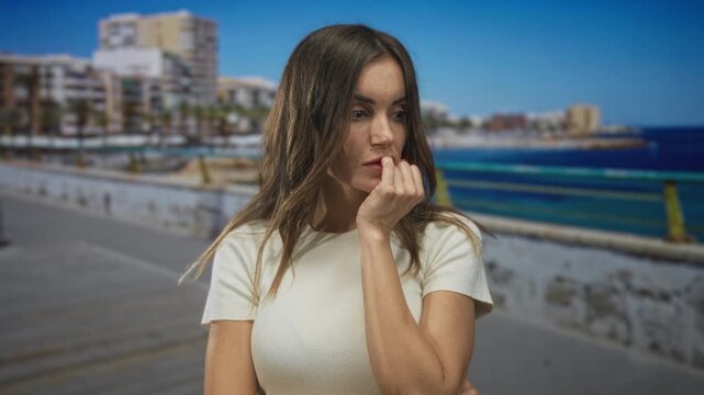 Young hispanic woman gazes sideways with finger to lips and bare arms crossed on seaside street; anxiety.