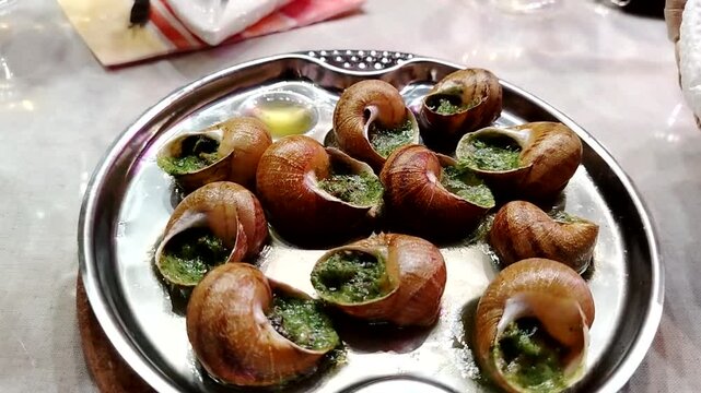 Plate of cooked escargot with garlic herb butter arranged in a circular pattern, garnished with olive oil, served on a silver platter in a dining setting