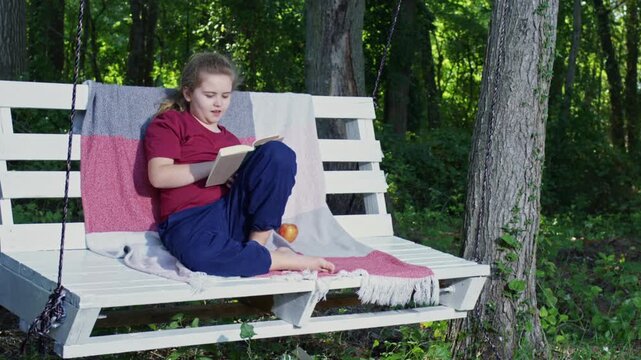 Reading supports childhood imagination. Child enjoys education in nature. Boy reads book while swinging slowly. Kid studies and relaxes outside. Child reading shows love for literature.