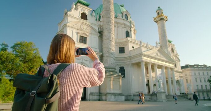Young woman traveler seen from behind taking photos with her smartphone of the historic Karlskirche church in Vienna, Austria. High-quality video for travel and lifestyle projects in summer