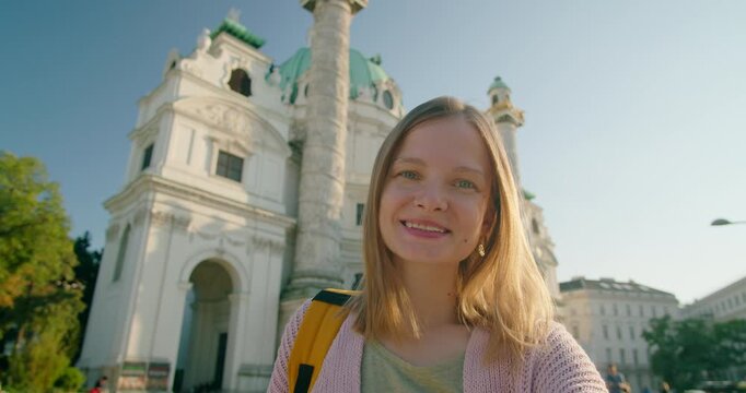 Young Caucasian woman traveler or blogger smiles and waves while making a selfie video call or recording a vlog in front of the historic Karlskirche in Vienna, Austria, on a sunny summer day