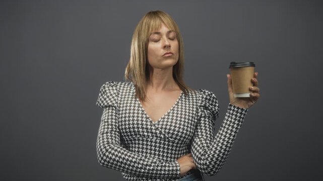Woman holds disposable coffee cup in right hand, arms crossed, looking at cup in studio; skepticism appraisal.