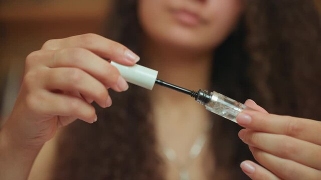 Closeup hands applying mascara in hotel room, curlyhaired woman preparing for evening, wand pulled from clear tube, lashes coated with clear serum, soft warm lighting and blurred background convey