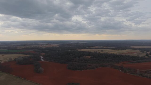 Drone aerial orbit over red dirt farmland and winding creek in rural Oklahoma under dramatic pre-storm clouds across the American Midwest countryside.