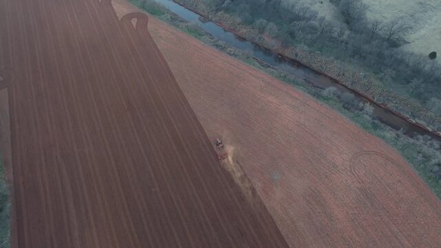 Drone aerial pulling back and tilting up revealing a tractor plowing red dirt farmland beside a winding creek in rural Oklahoma agricultural landscape.
