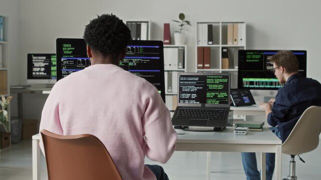 Rear view of young adult African American male coder sitting down at programming workstation then writing code on laptop and desktop computer next to colleagues workspace in modern office
