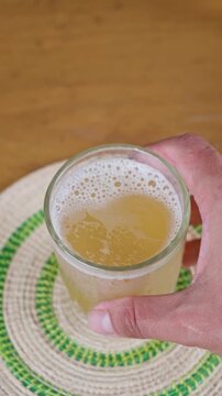 Hand holding clear glass of golden kombucha fermented probiotic beverage with foam bubbles close view on wooden table. 