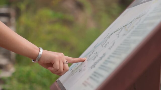 Close up hand points at map with fingertip, silver bracelet visible, detailed map text and route lines in focus, outdoor greenery blurred in background, careful map reading and route confirmation