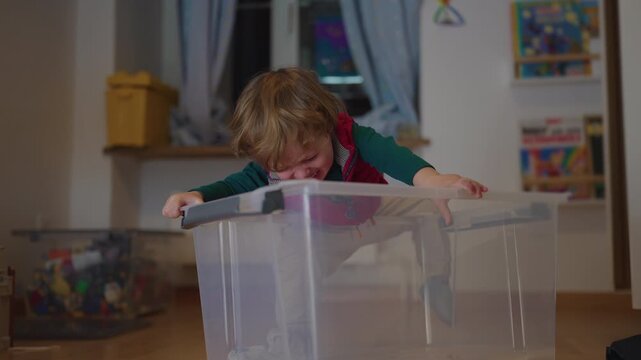 Frustrated toddler struggling to climb into large plastic storage box, determined childhood moment of effort, curiosity and playful persistence at home