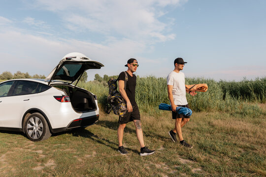 Two friends walking from a white car carrying chairs for a summer road trip in the countryside