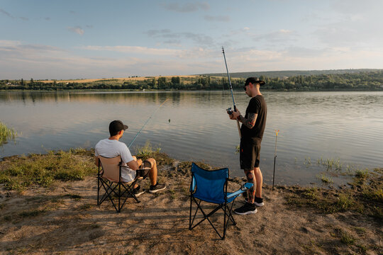 Two friends fishing on a peaceful lakeshore at sunset with folding chairs and rods