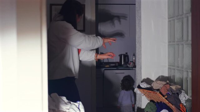 Mother creating shadow figures with hands on kitchen wall to entertain toddler, playful family bonding moment in cozy apartment hallway