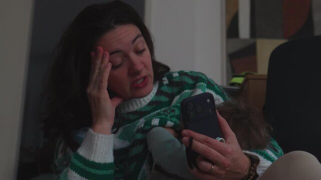 Mother breastfeeding toddler while holding smartphone at home, multitasking parent speaking on phone during intimate family moment