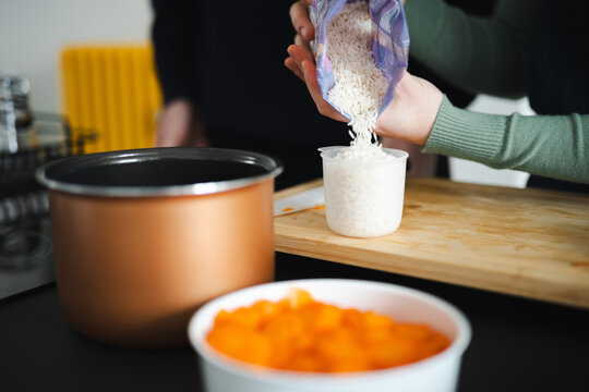 A person pours uncooked rice from a bag into a measuring cup, preparing to cook a meal with chopped vegetables and a pot on the stove.
