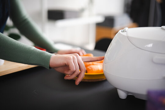 A person's hand presses a button on a white kitchen appliance, with carrots in the background.