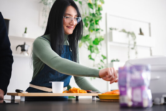A woman in an apron prepares food in a bright kitchen, chopping vegetables on a wooden board.