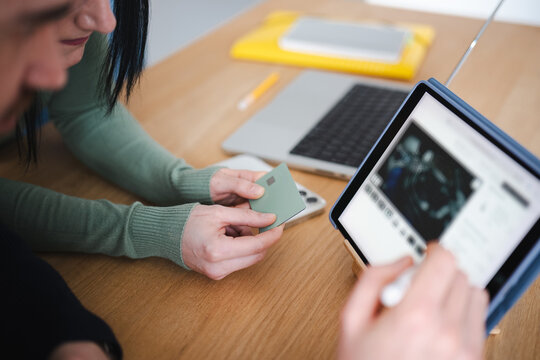 Two people are using a tablet and a credit card to make a purchase online, with a laptop and other items on the desk.