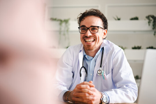 A smiling doctor in a white coat with a stethoscope listens attentively to a patient during a consultation in a bright, modern office.