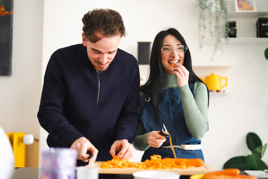 A man and woman prepare vegetables together in a kitchen. The woman smiles as she eats a piece of carrot while the man slices more carrots on a cutting board.