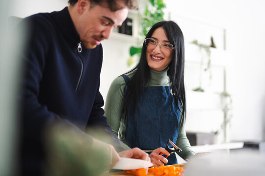 A man and a woman are preparing food together in a kitchen. The woman smiles as she holds a peeler over chopped carrots.
