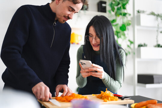 A man slices carrots while a woman looks at her phone in a kitchen. They are preparing food together.
