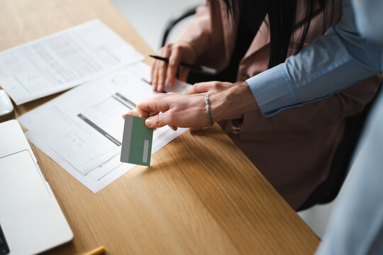 A person holds a payment card over documents on a desk, with another person's hand nearby, suggesting a business transaction or financial review.
