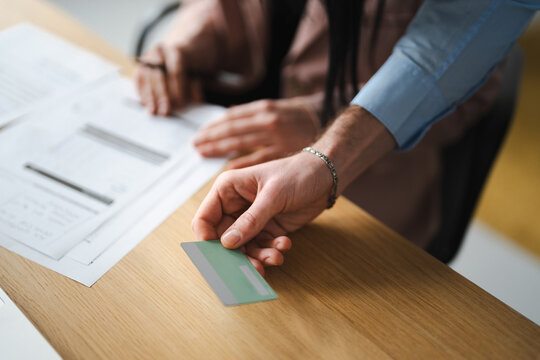 A man's hand offers a credit card to a woman who is reviewing documents at a desk, suggesting a transaction or payment.