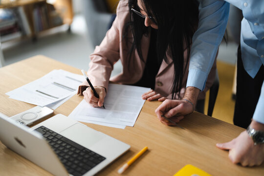 A woman with glasses signs a document at a desk while a man in a blue shirt leans in to observe.